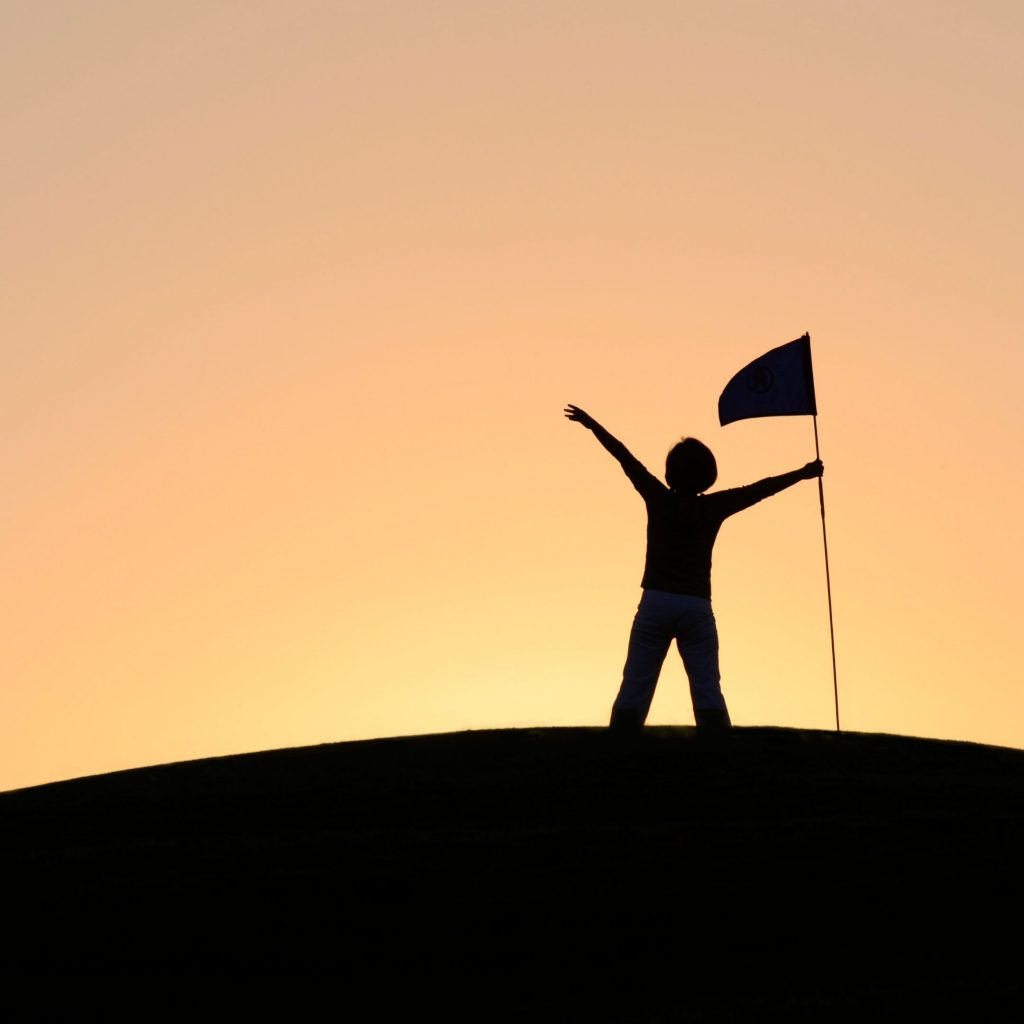 "Silhouette of a person raising their arms in victory while holding a flag on a hill against a sunset background, representing success and accomplishment."