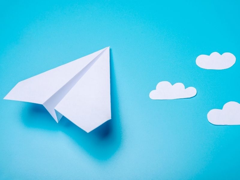 "A white paper airplane on a blue background with paper cut-out clouds, representing creativity, travel, and aspirations."