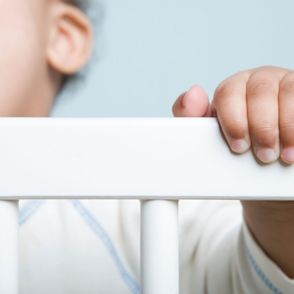 "A baby's small hand grasping the white crib railing, with a soft-focus background highlighting the child's gentle features."