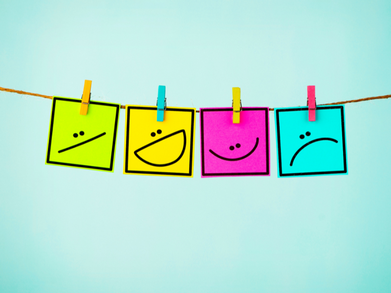 "Four sticky notes in bright colors clipped to a string, each featuring a unique facial expression symbolizing different emotions."