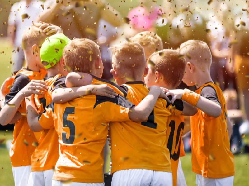 "Kids' soccer team in orange jerseys embracing in a celebratory huddle with confetti in the air, representing teamwork and success."