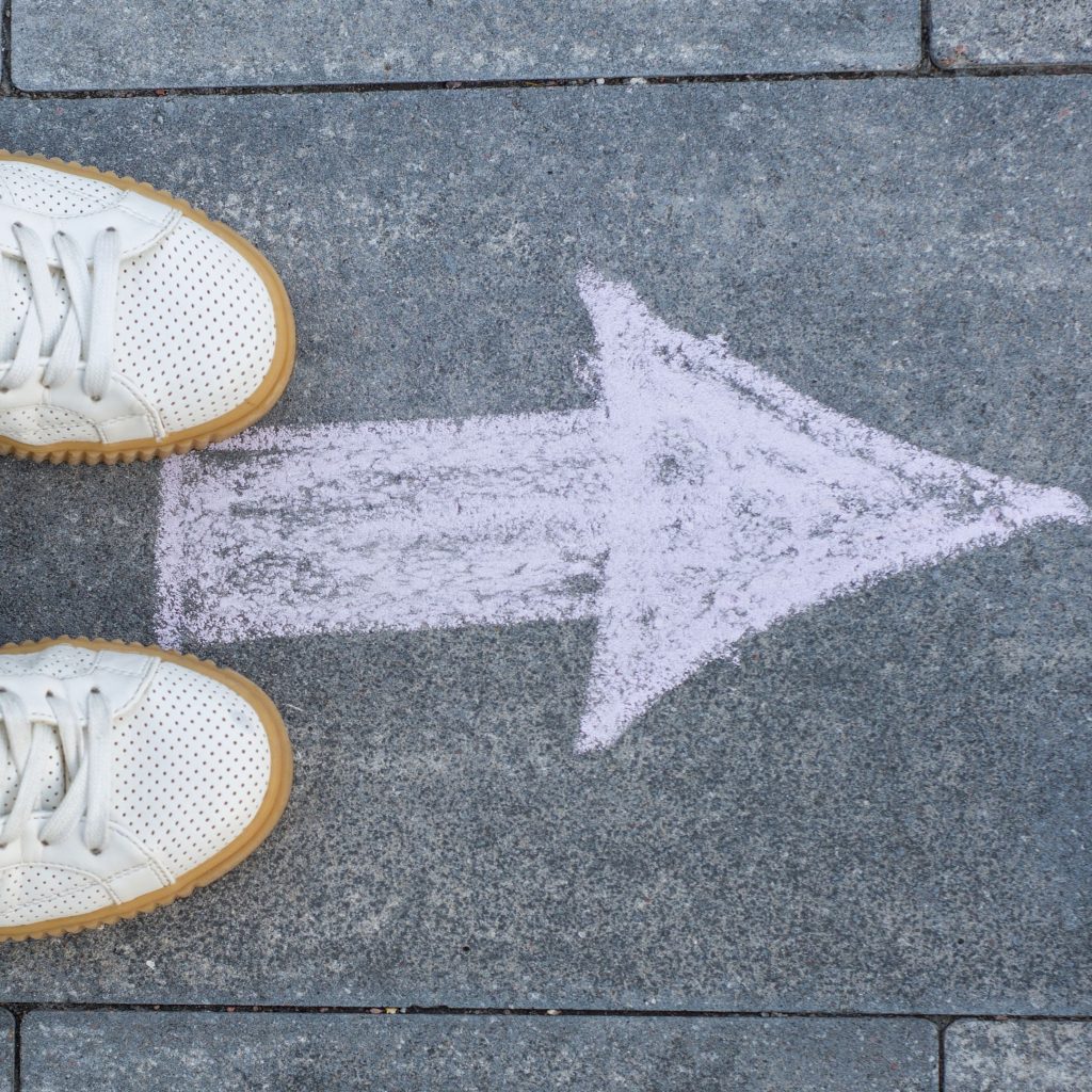 "White sneakers on a stone pavement with a large chalk arrow pointing forward, representing direction and progress."